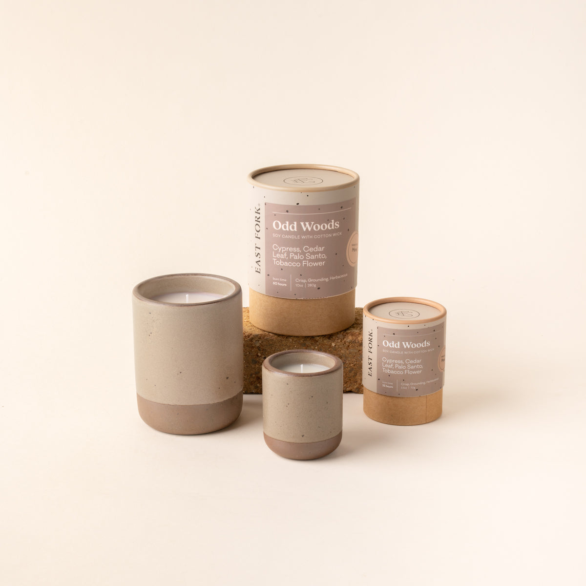 Small and large ceramic vessel in a warm pale brown color with candles inside, arranged with tube packaging that reads 'Odd Woods'
