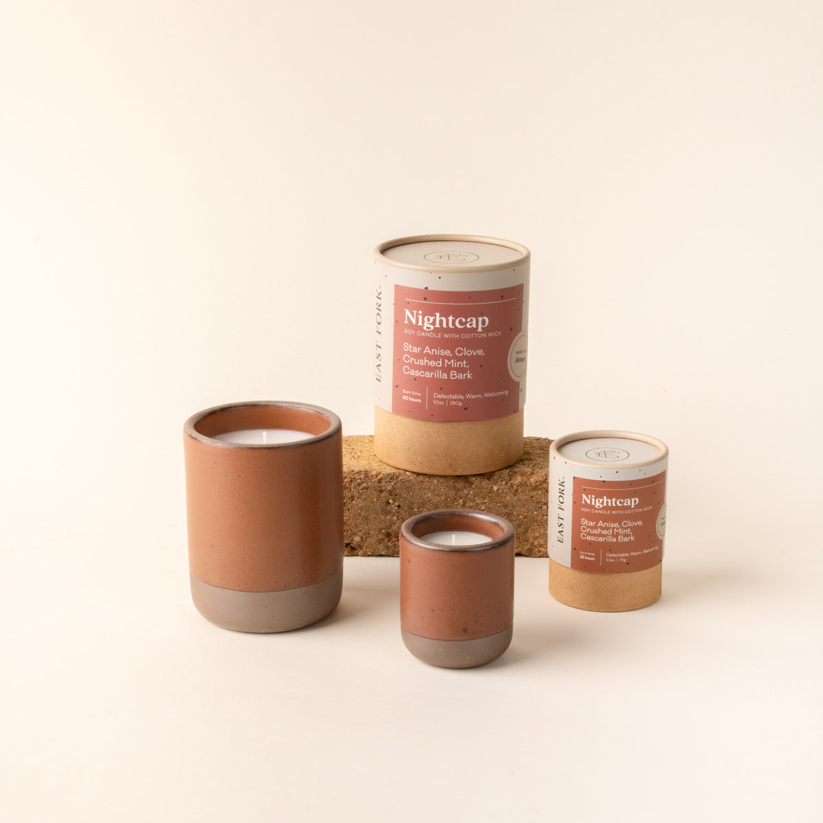 Small and large ceramic vessel in a cool terracotta color with candles inside, arranged with tube packaging that reads 'Nightcap'