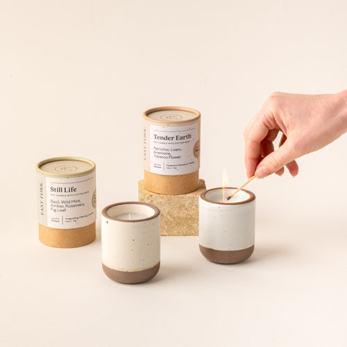 Two small ceramic vessels in cream off-white and cool white colors with candles inside each are gathered next to their cardboard packaging tubes. A hand is lighting the cool white candle with a match.