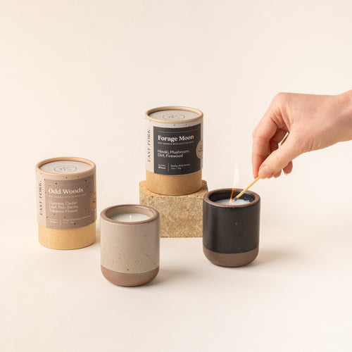 Two small ceramic vessels in pale warm brown and graphite black colors with candles inside each are gathered next to their cardboard packaging tubes. A hand is lighting the cool white candle with a match.