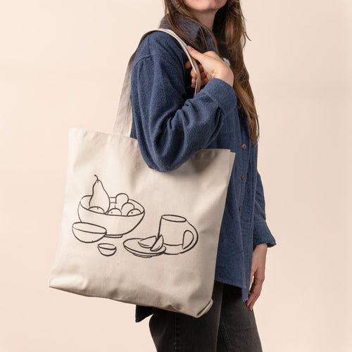 A person holding a beige cotton tote bag with a black printed drawing of a bowl and a mug.