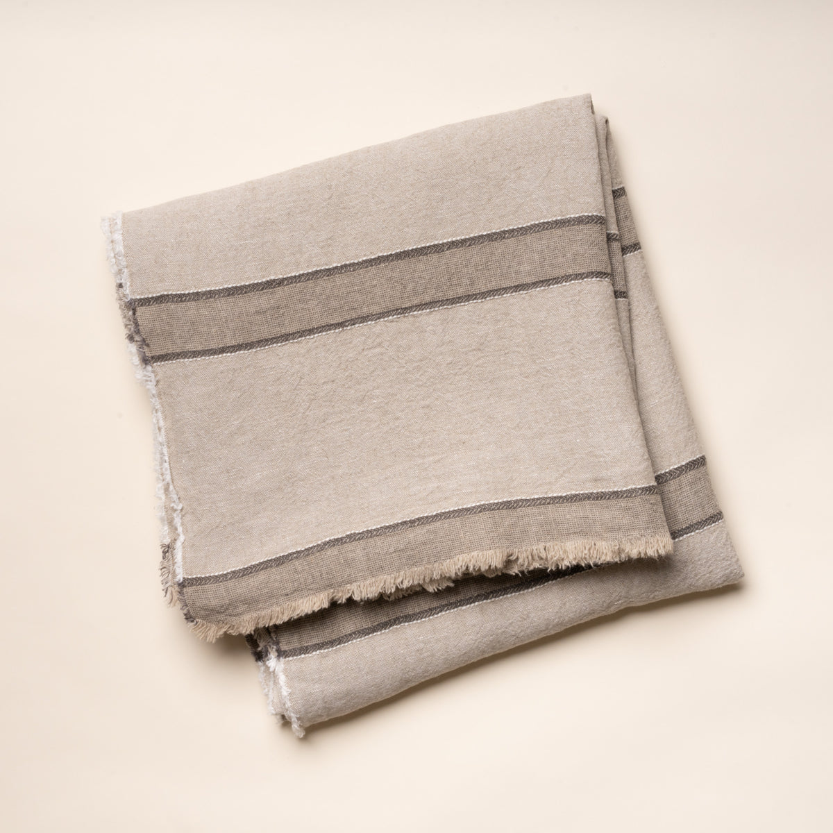 Neutral-toned linen tablecloth with woven gray stripes and fringed edges, neatly folded on a clean light background.