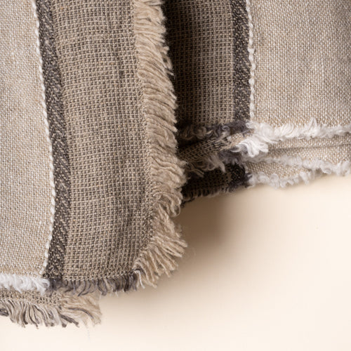 Close up of a neutral-toned linen tablecloth with woven gray stripes and fringed edges, neatly folded on a clean light background.