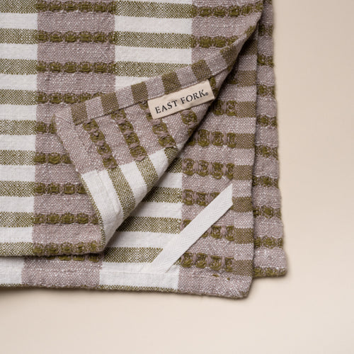 Corner of a folded woven kitchen towel with olive green and beige striped pattern with a tag that reads 'East Fork' and a white loop for hanging on a light background.