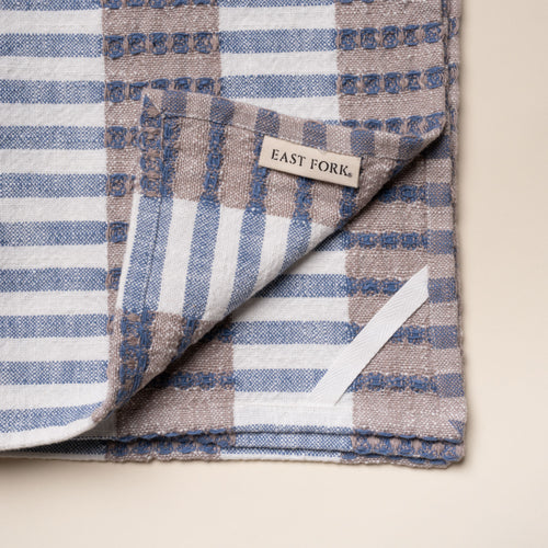 Corner of a folded woven kitchen towel with blue and beige striped pattern with a tag that reads 'East Fork' and a white loop for hanging on a light background.