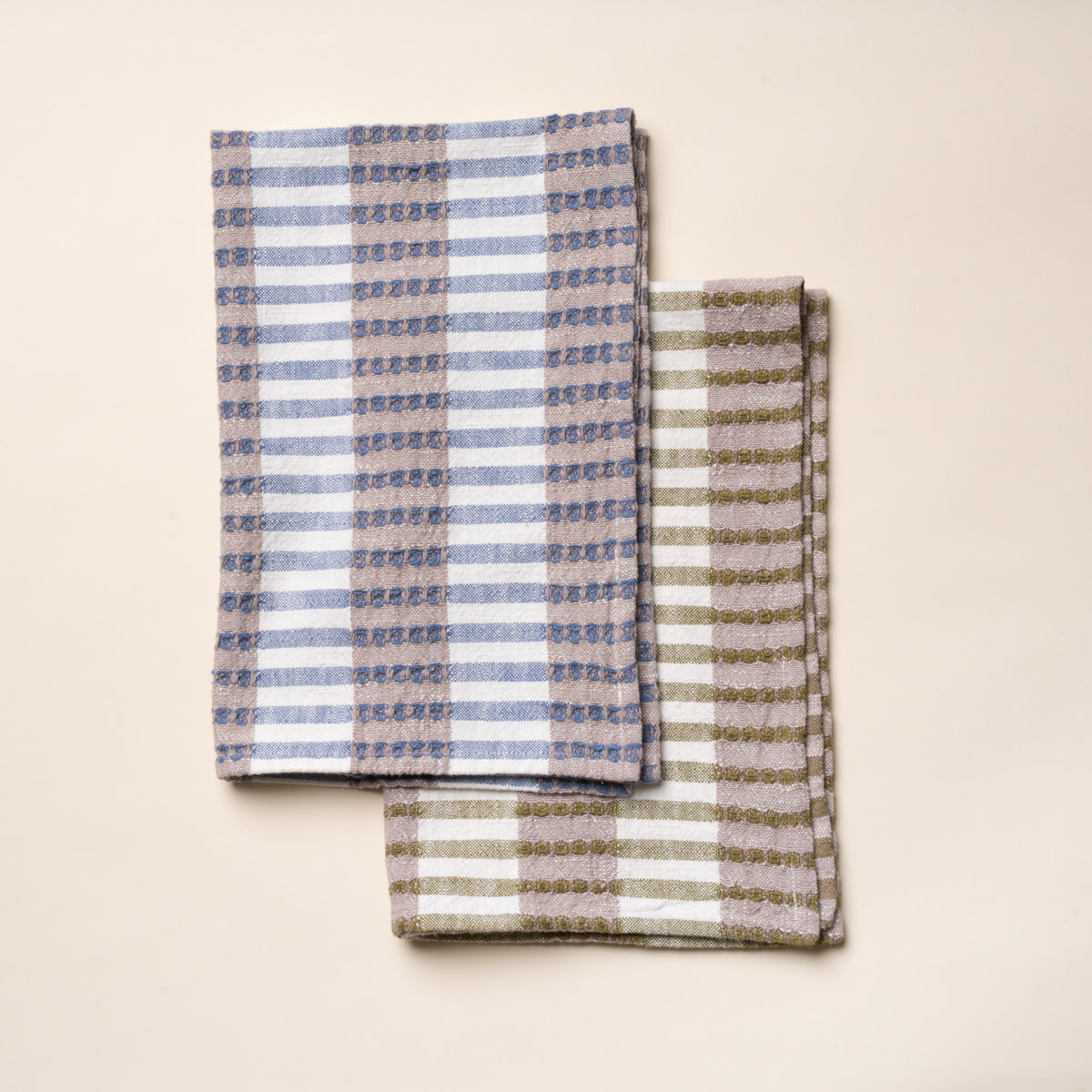 Two different folded woven kitchen towels - one in blue and beige and one in olive and beige - with a striped pattern on a light background.