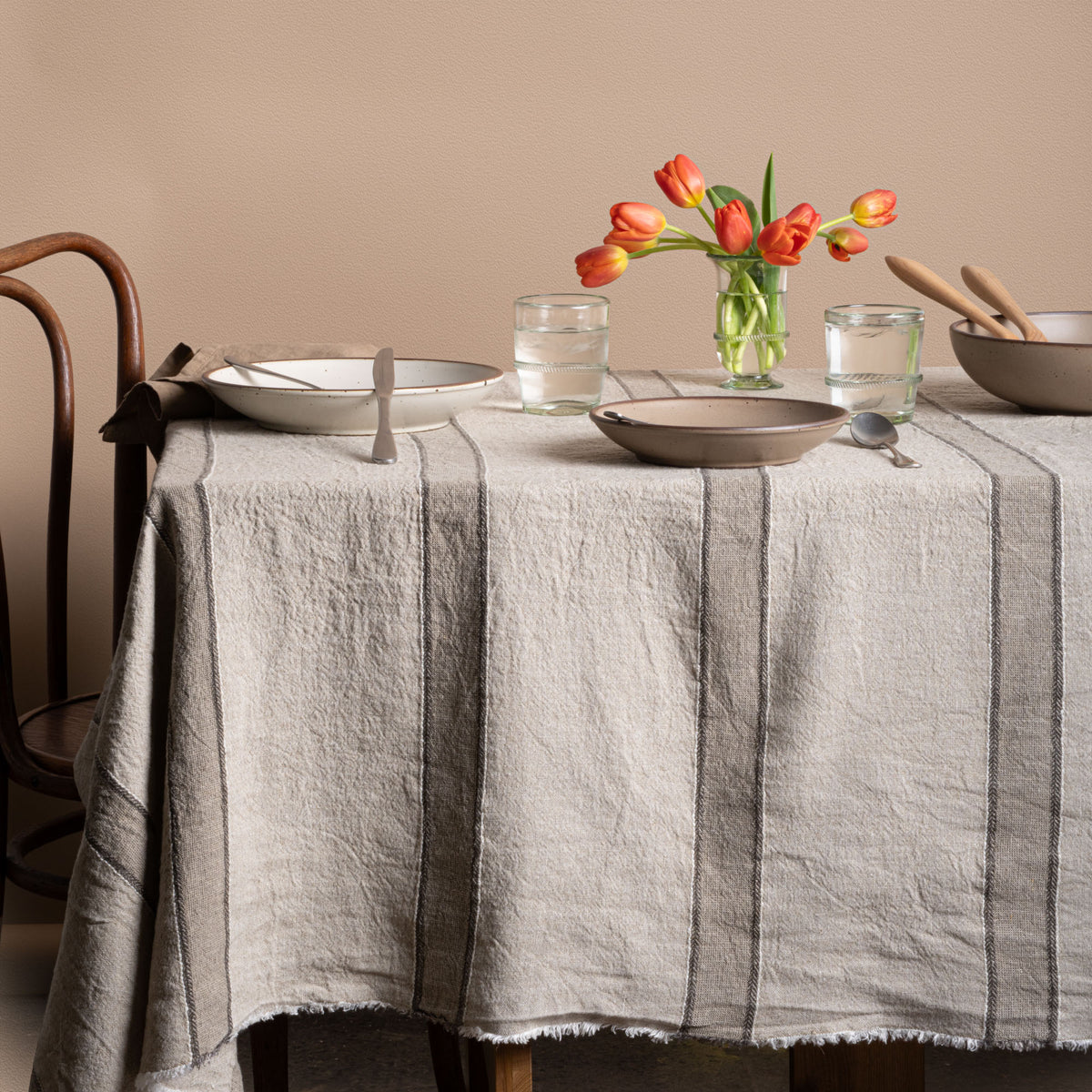 Rustic dining table styled with a neutral striped linen tablecloth, ceramic dinnerware, banded glasses and a vase of tulips.