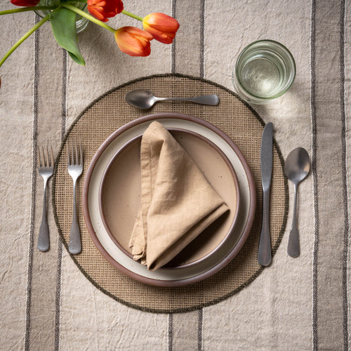Dining table setting with plates, cutlery, and a napkin on a round woven placemat and striped tablecloth.