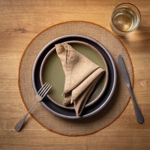 Dinner setting with plates, napkin, fork, knife, and glass with a round woven placemat with a sewn rust rim on a wooden table.