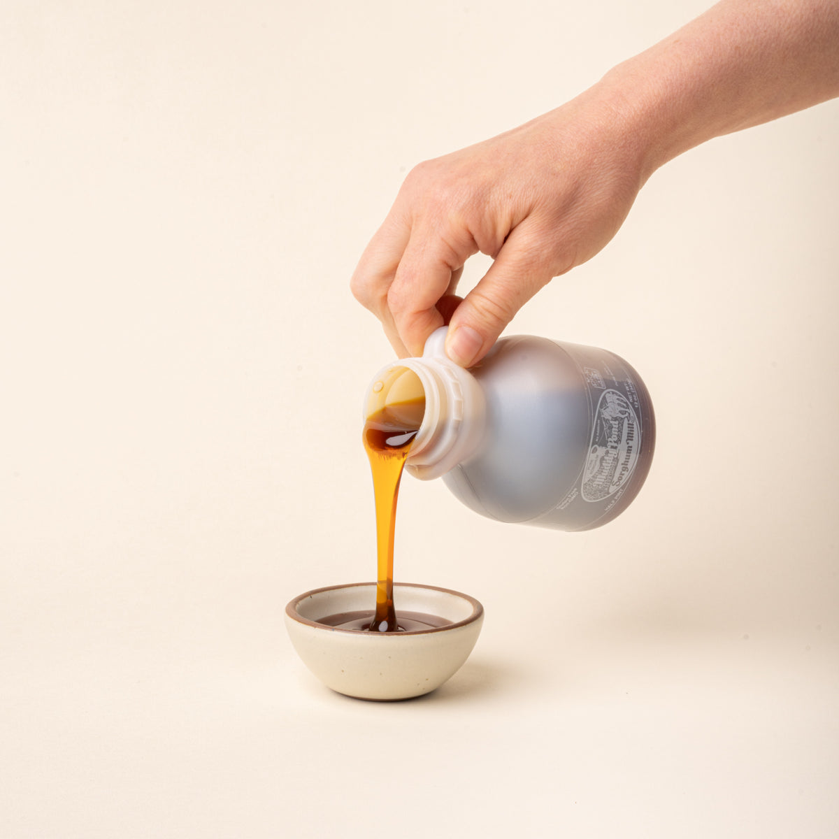 Hand pours amber sorghum syrup from a bottle into a small ceramic bowl on a plain white background.