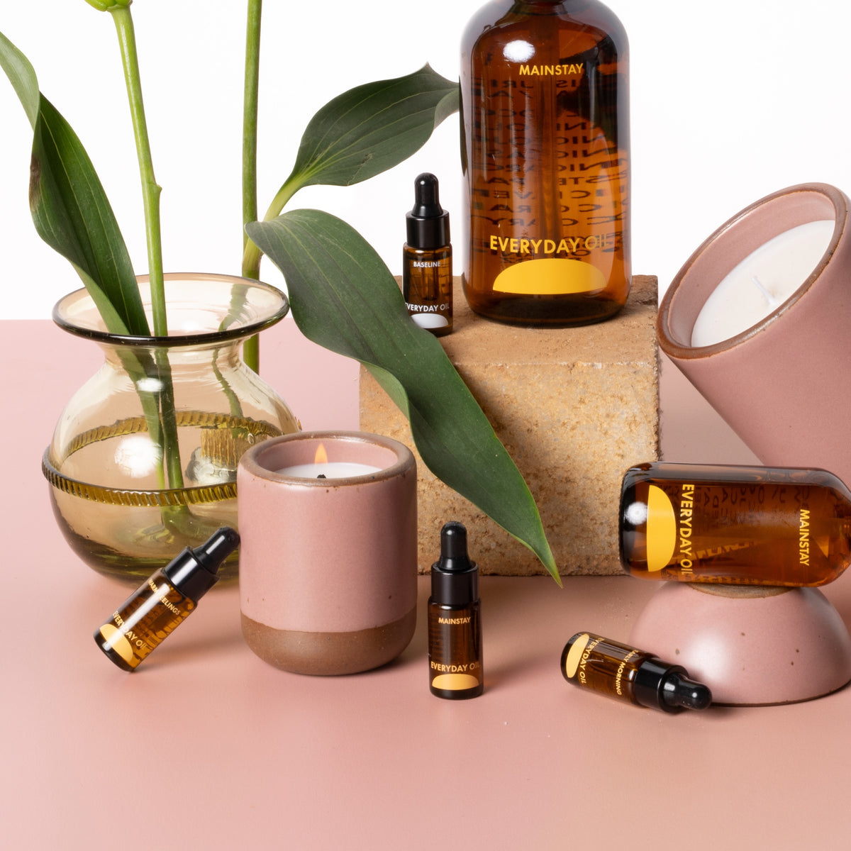 Styled still life of amber glass bottles of body oil with droppers, a leafy plant in a glass vase, and ceramic candle holders on a soft pink surface.