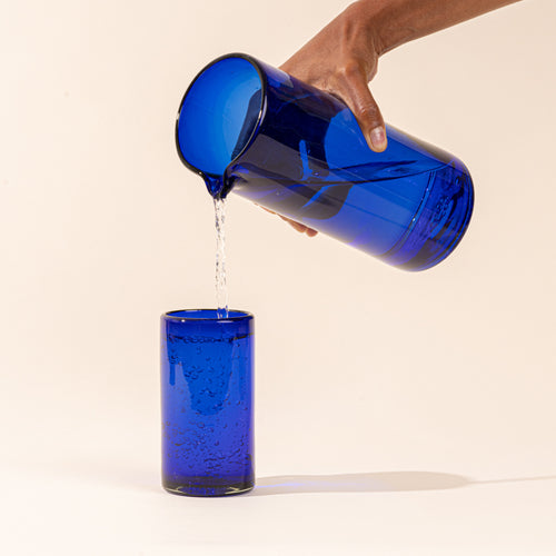 A hand holds a cobalt blue tall transparent glass pitcher with small spout at the top, pouring water into a matching tumbler.