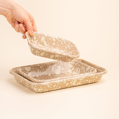 A hand placing a small dish into a nesting trio rectangular baking dishes in a neutral tan color with off-white splattered pattern