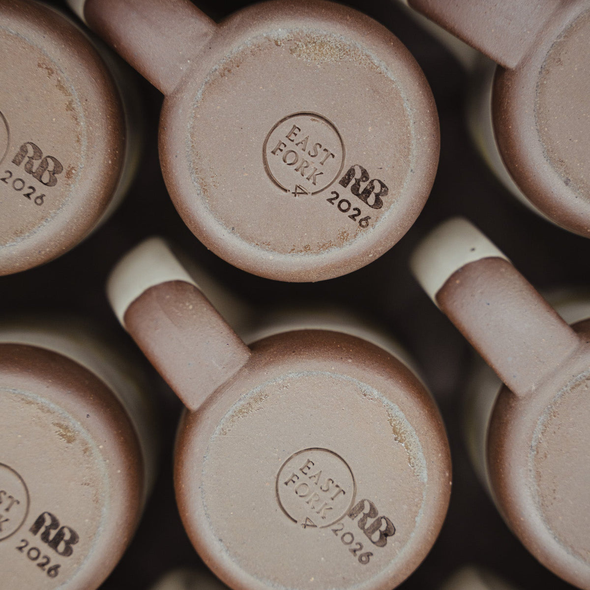 Close-up of multiple ceramic mugs arranged in a grid, showing the bottom of each mug with a stamped logo reading 'EAST FORK RB 2026'. 