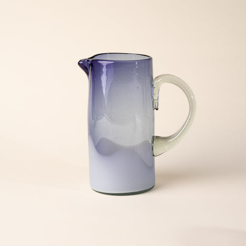 White and clear iris handblown glass pitcher with organic wave design with clear handle, casting a soft shadow on a beige backdrop.