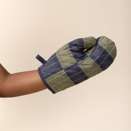 A hand wears a quilted oven mitt with green and navy patchwork pattern and white stitched lines.