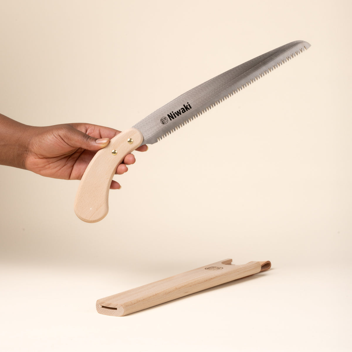 A hand holds a Niwaki wooden-handled pruning saw with protective wooden sheath.