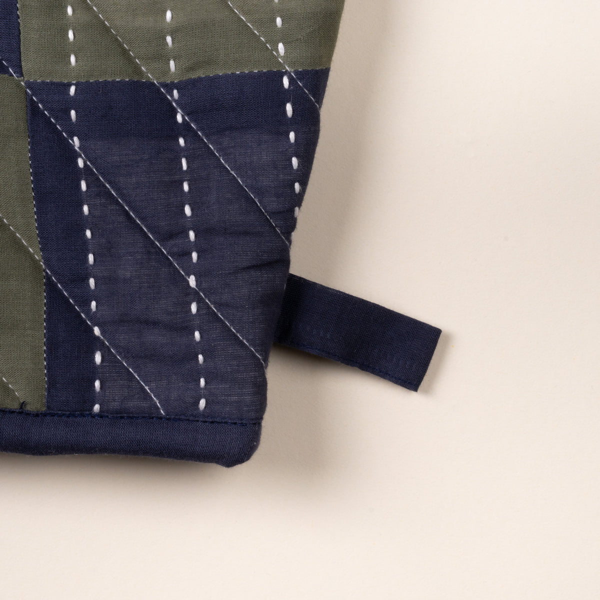 Close up of a corner of a quilted oven mitt with green and navy patchwork pattern and white stitched lines, shown on a neutral background.