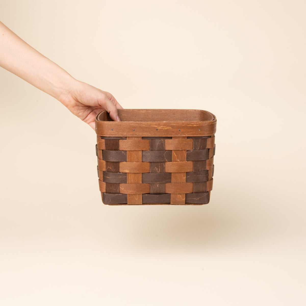 Hand holds out a medium rectangular woven wood basket in two-tone brown with an open top and reinforced rim, shown against a neutral background.