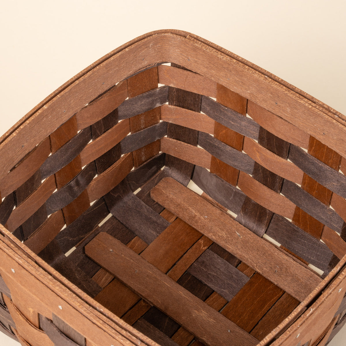 Closeup of the inside of a medium rectangular woven wood basket in two-tone brown with an open top and reinforced rim, shown against a neutral background.