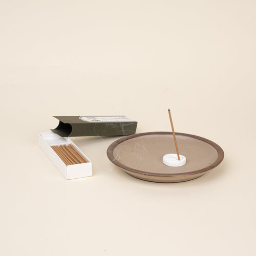 Lit stick incense in a holder on a ceramic dish, with open box of incense sticks beside it.