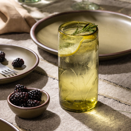 Olive etched glass tumbler filled with ice, lemon slices, and a rosemary sprig beside a bowl of blackberries and ceramic dinnerware.