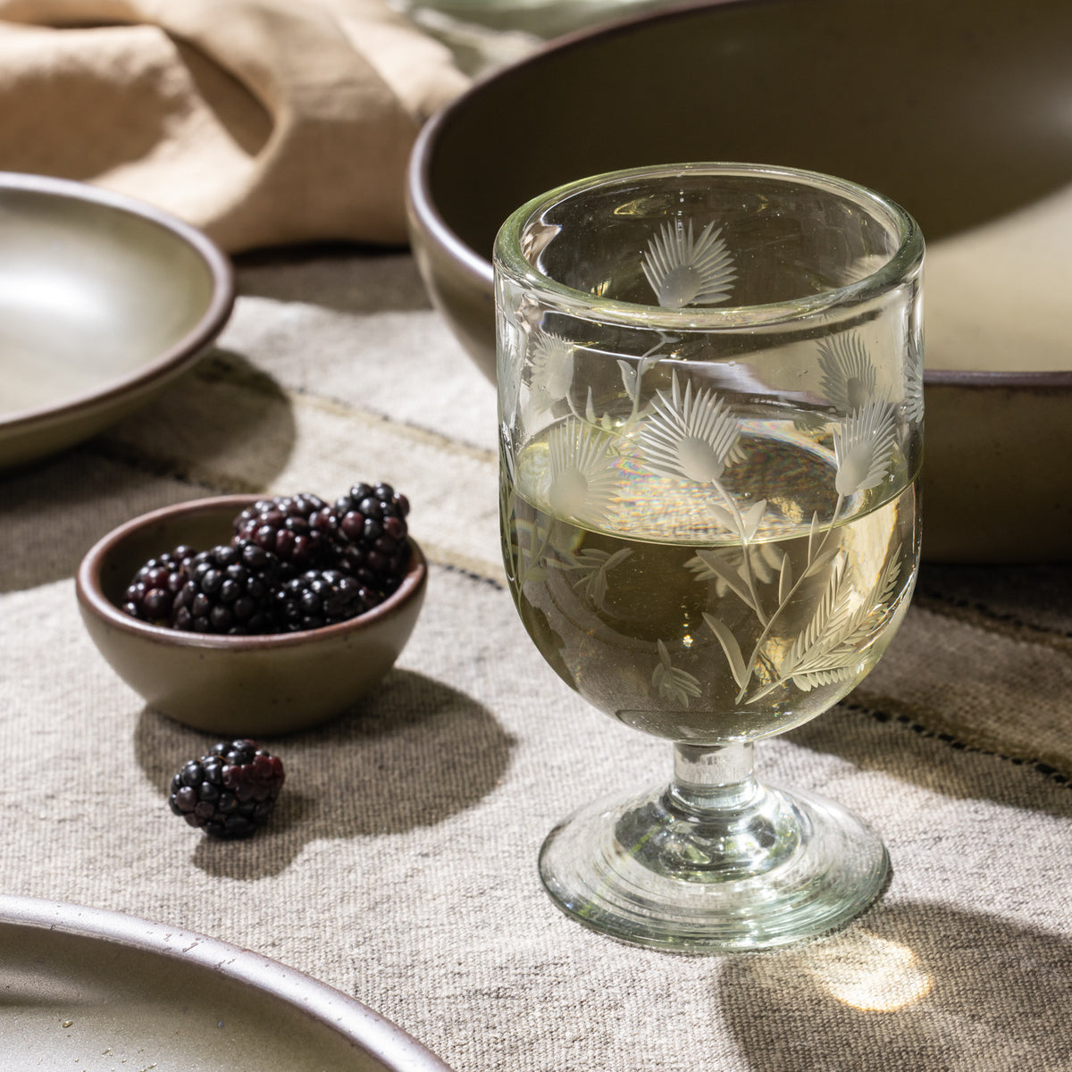 Etched stemmed glass filled with a clear beverage beside a small bowl of blackberries and ceramic dinnerware.