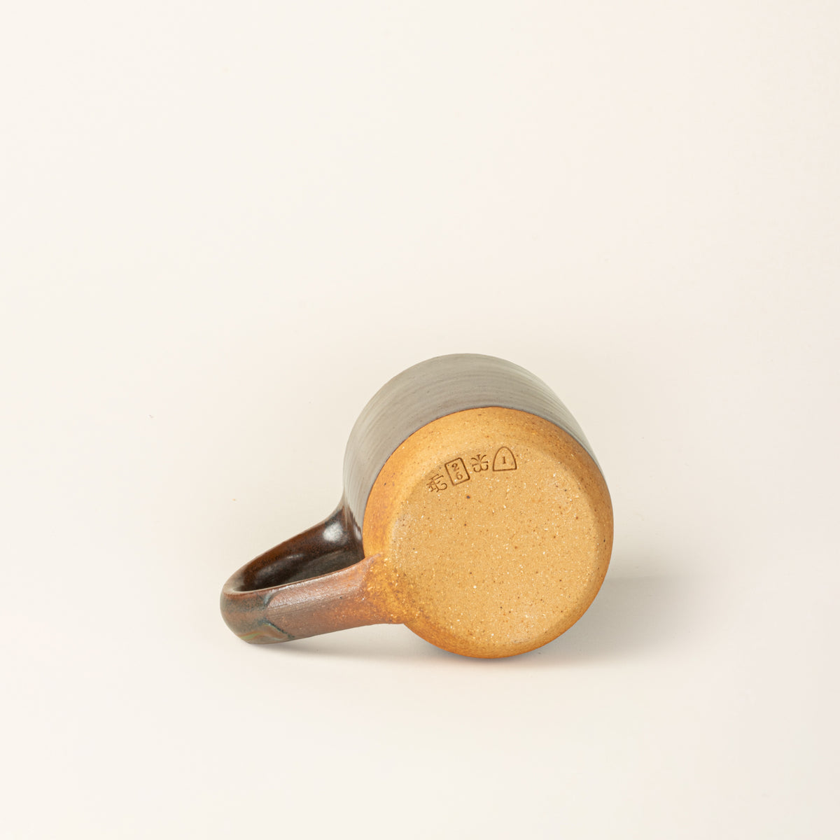 Bottom of a handmade ceramic mug with a wood fired light earthy green glaze with an unglazed base, showing off 4 stamps.