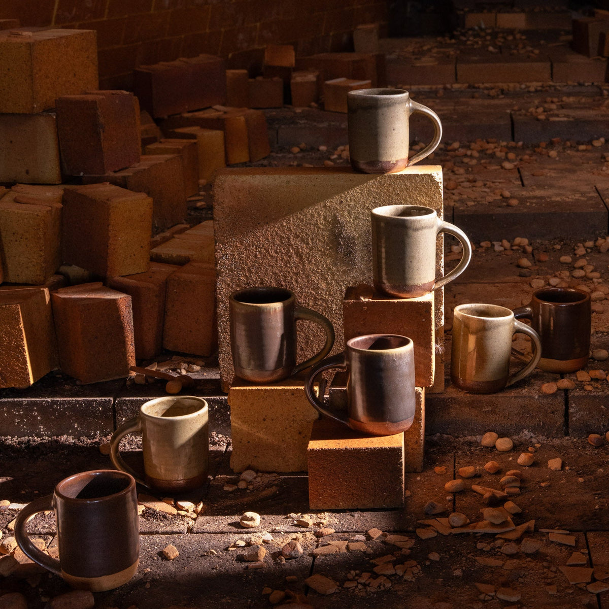 A group of handmade ceramic mugs in various earthy glazes, styled on and around rustic clay bricks in a wood fired kiln.