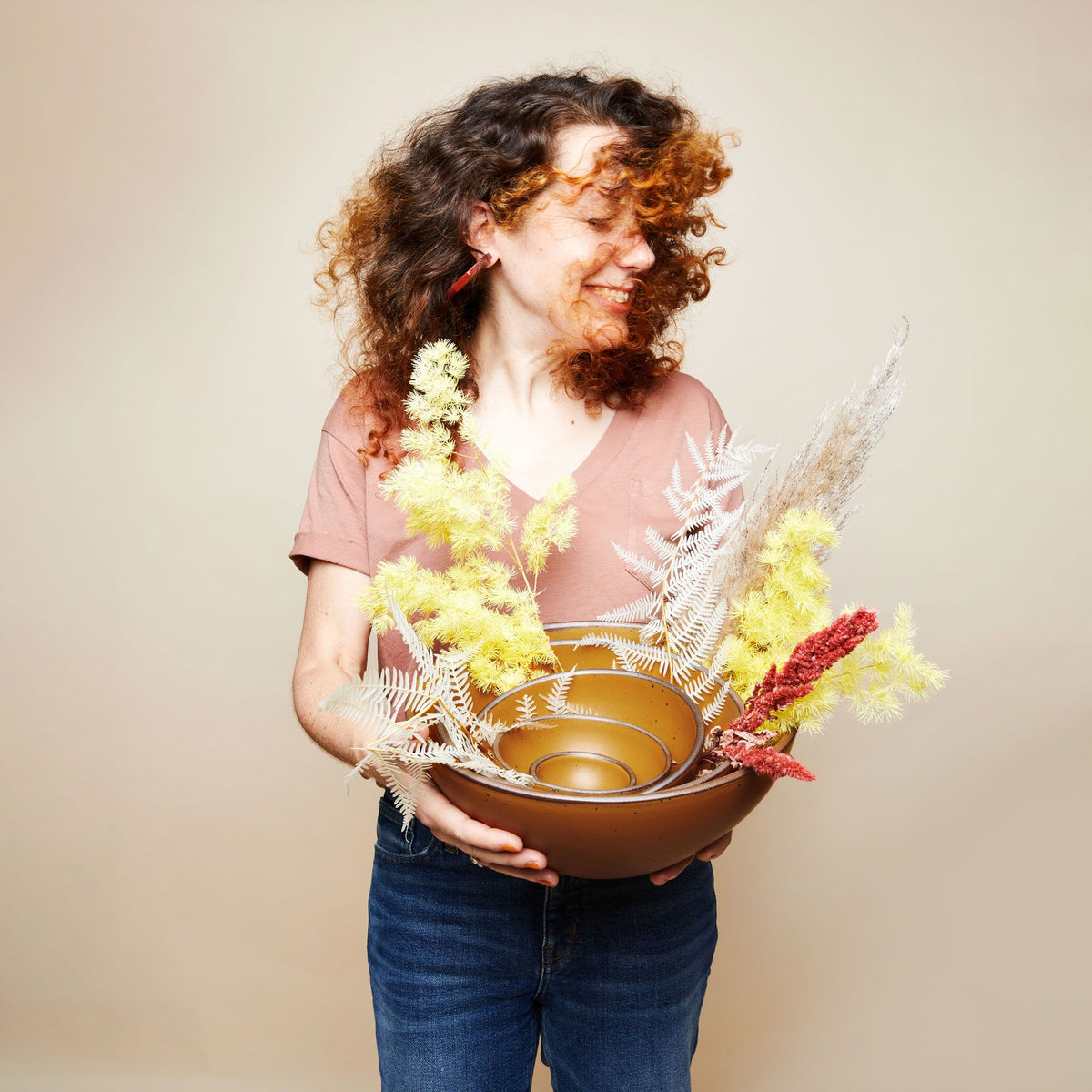 A person is smiling and flipping their hair as they hold a stack of nesting bowls in a dark gold color with dried grasses and florals.