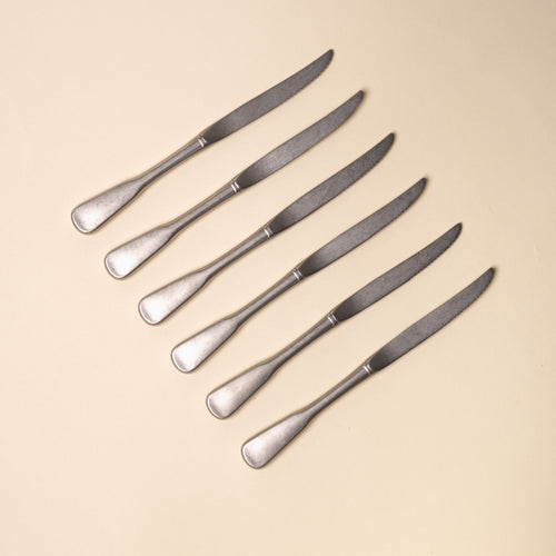 Six matte stainless steel dinner steak knives with rounded handles arranged diagonally on a neutral beige background.