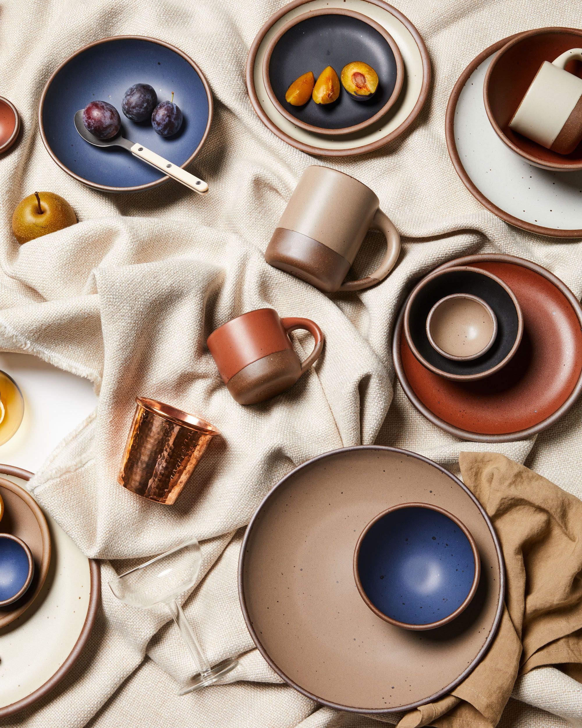 An overhead view of artfully arranged ceramic plates, bowls, and mugs in various sizes and colors with fruits, copper cup, and fabrics