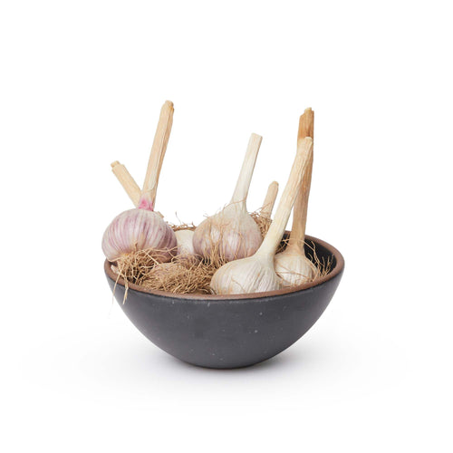 A medium rounded ceramic bowl in a graphite black color featuring iron speckles and an unglazed rim, filled with garlic