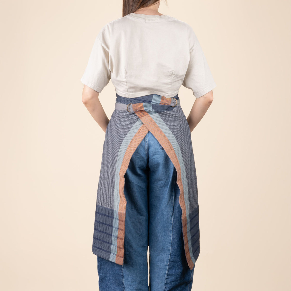A view of the back of a person wearing a woven kitchen waist apron in navy and slate blue with light blue and rust stripe accents with a front pocket and long tie straps.