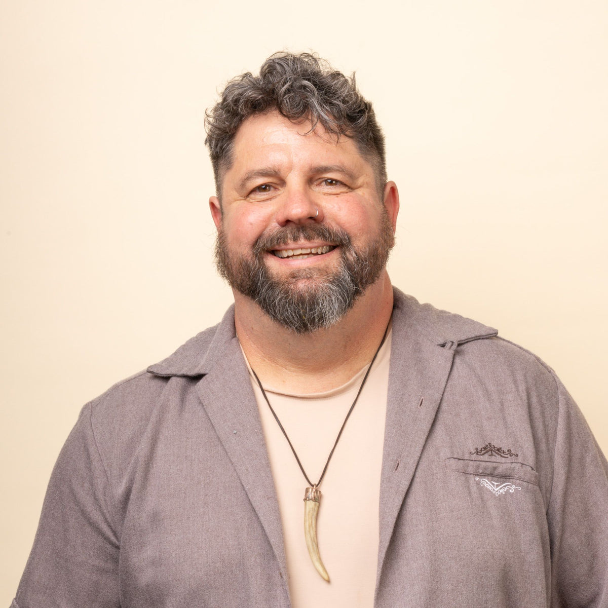 A person with a beard and curly hair is smiling, wearing a light beige shirt and a gray open-collared jacket, with a necklace featuring a long pendant. The background is a plain, soft beige color.