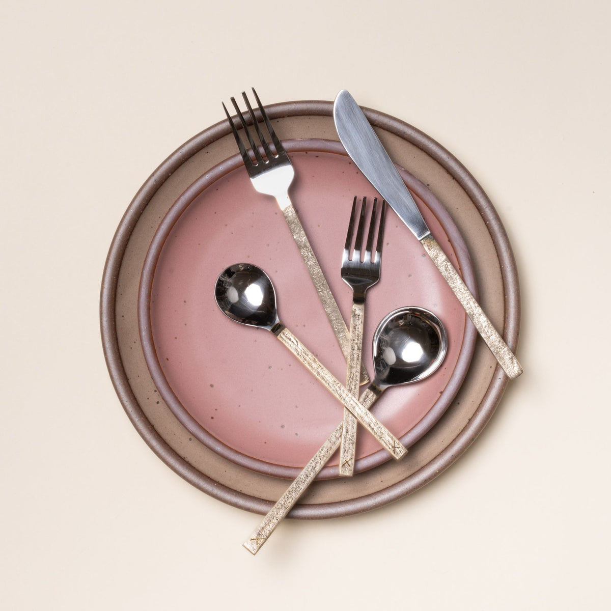 Five-piece stainless steel and brass flatware set with two forks, a knife, and two spoons artfully arranged on a pale brown dinner plate and dusty pink side plate.