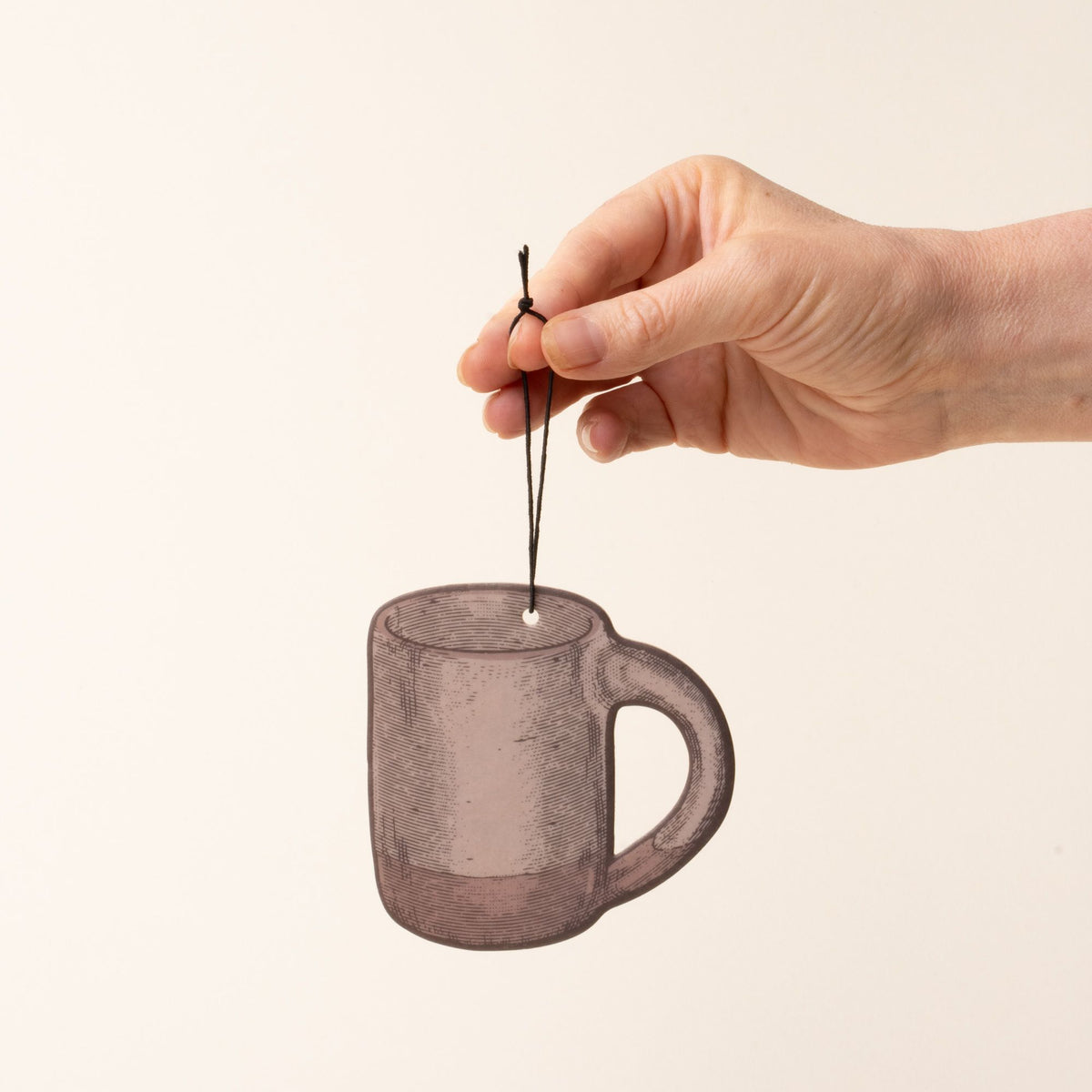 A hand holding an illustrated mug-shaped air freshener in a pale warm brown color with a black hanging string against a light cream background.