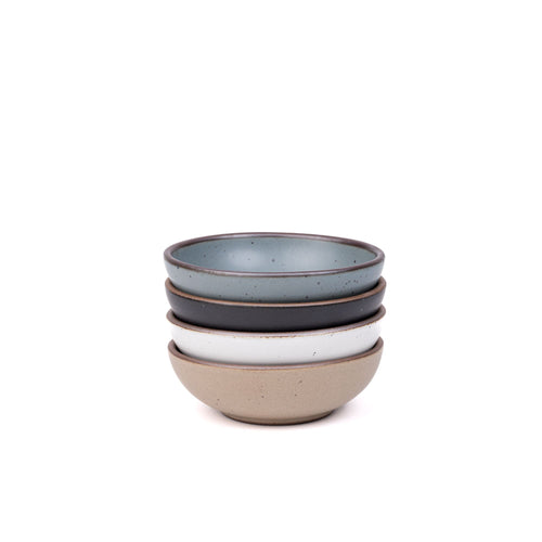 A stack of 4 small shallow ceramic bowls in a blue-grey, graphite black, cool white, and warm pale brown colors featuring iron speckles and an unglazed rim