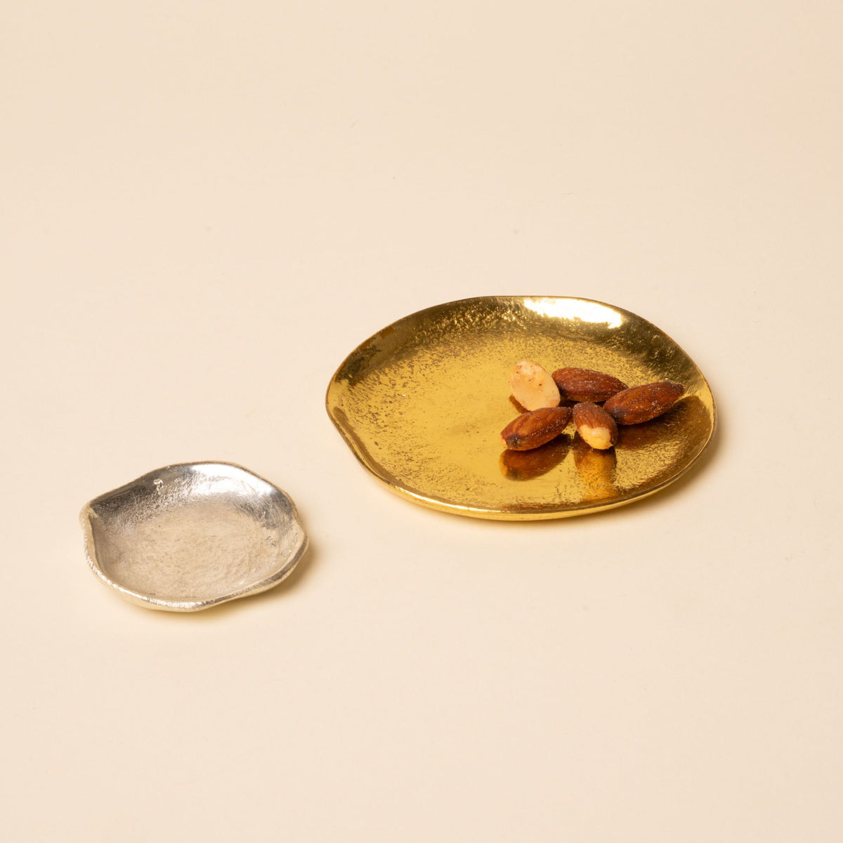 Two small hammered metal dishes, one gold with almonds and one silver, placed side by side on a neutral background.