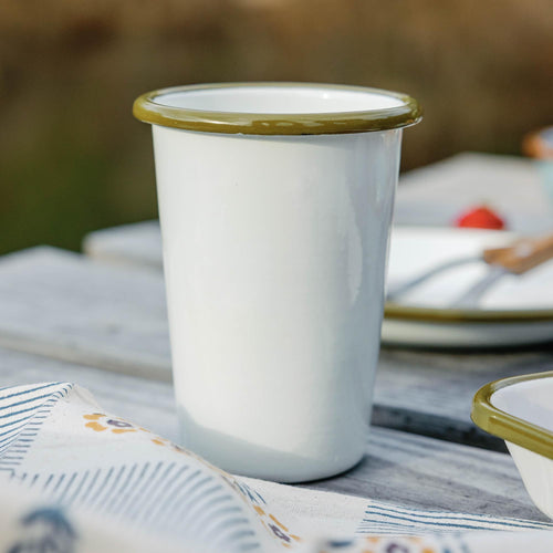 White enamel tumbler with an olive green rim sitting on a weathered wooden deck outdoors.