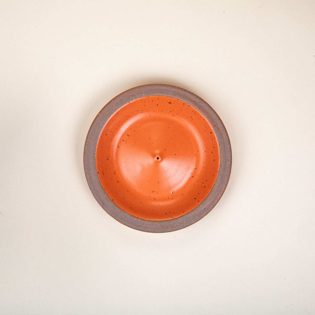An overhead view of a ceramic plate-shaped incense stick holder with an unglazed rim and glazed in a bold orange color featuring lots of iron speckles.