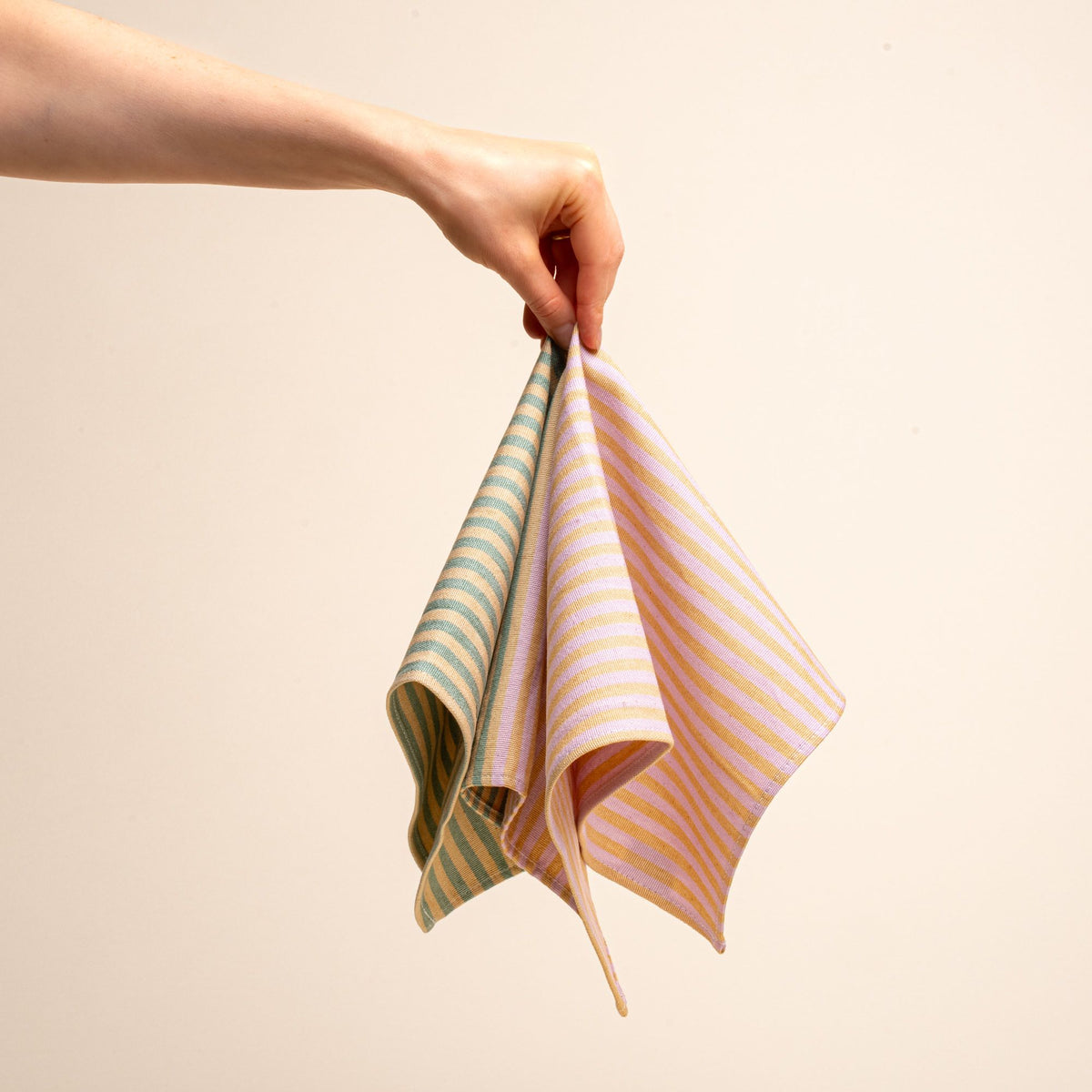 A hand holds a napkin featuring color blocks of striped peach and pink, and striped peach and green.