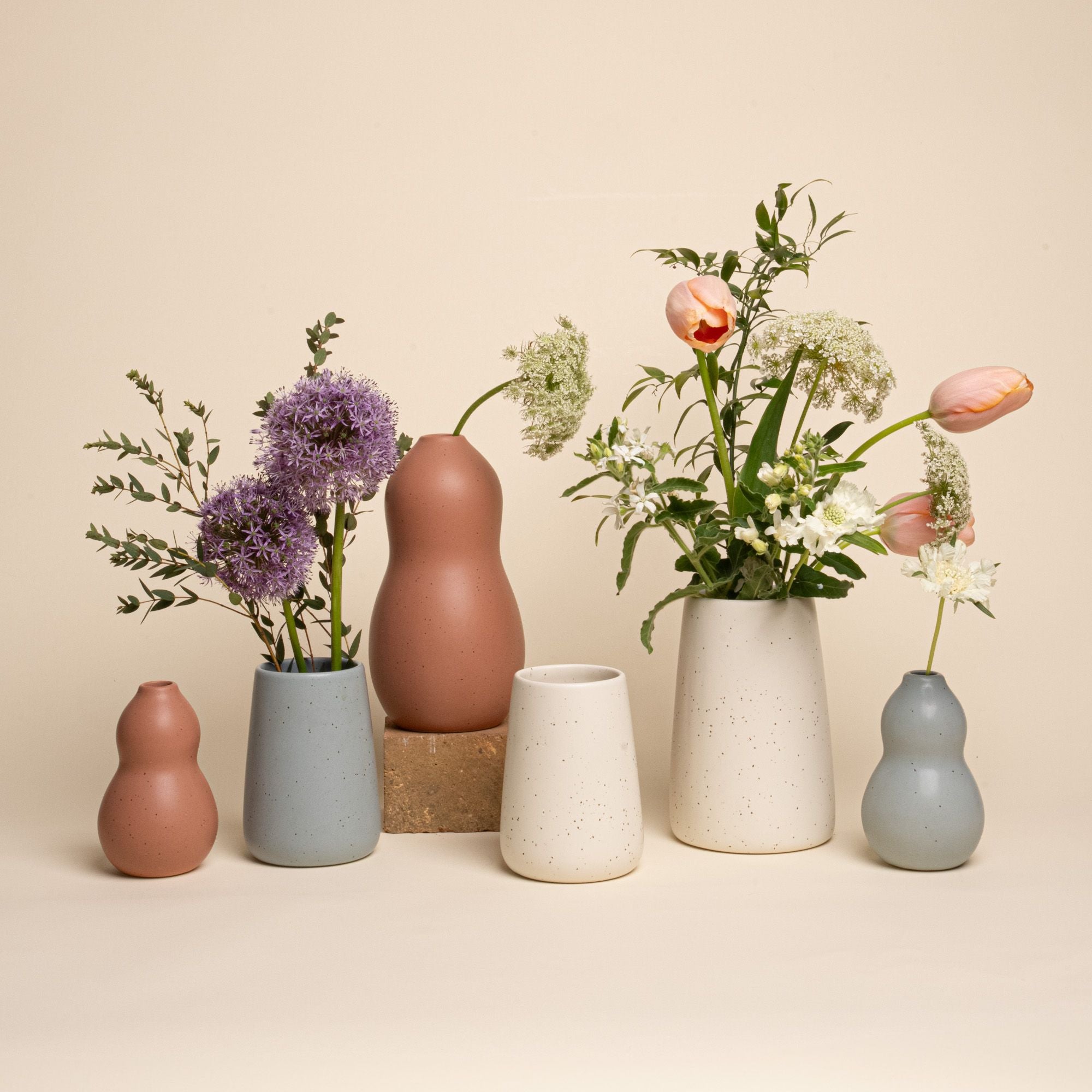 A group of six ceramic vases in terracotta, muted blue, and cream with speckling, each holding fresh flowers including tulips, alliums, and wildflowers, arranged on a neutral background.