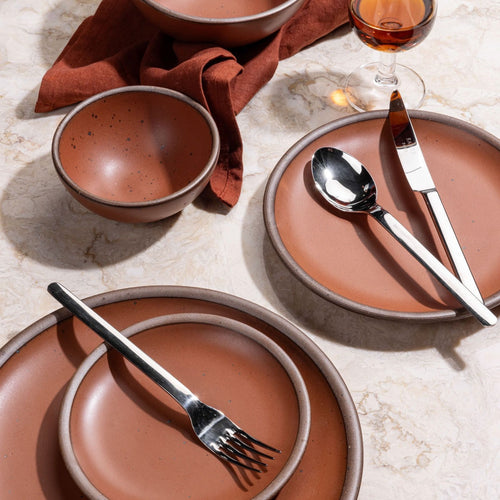A collection of terracotta-colored ceramic plates and bowls on a marbled surface, accompanied by stainless steel cutlery, a brown napkin, and a glass of amber liquid.