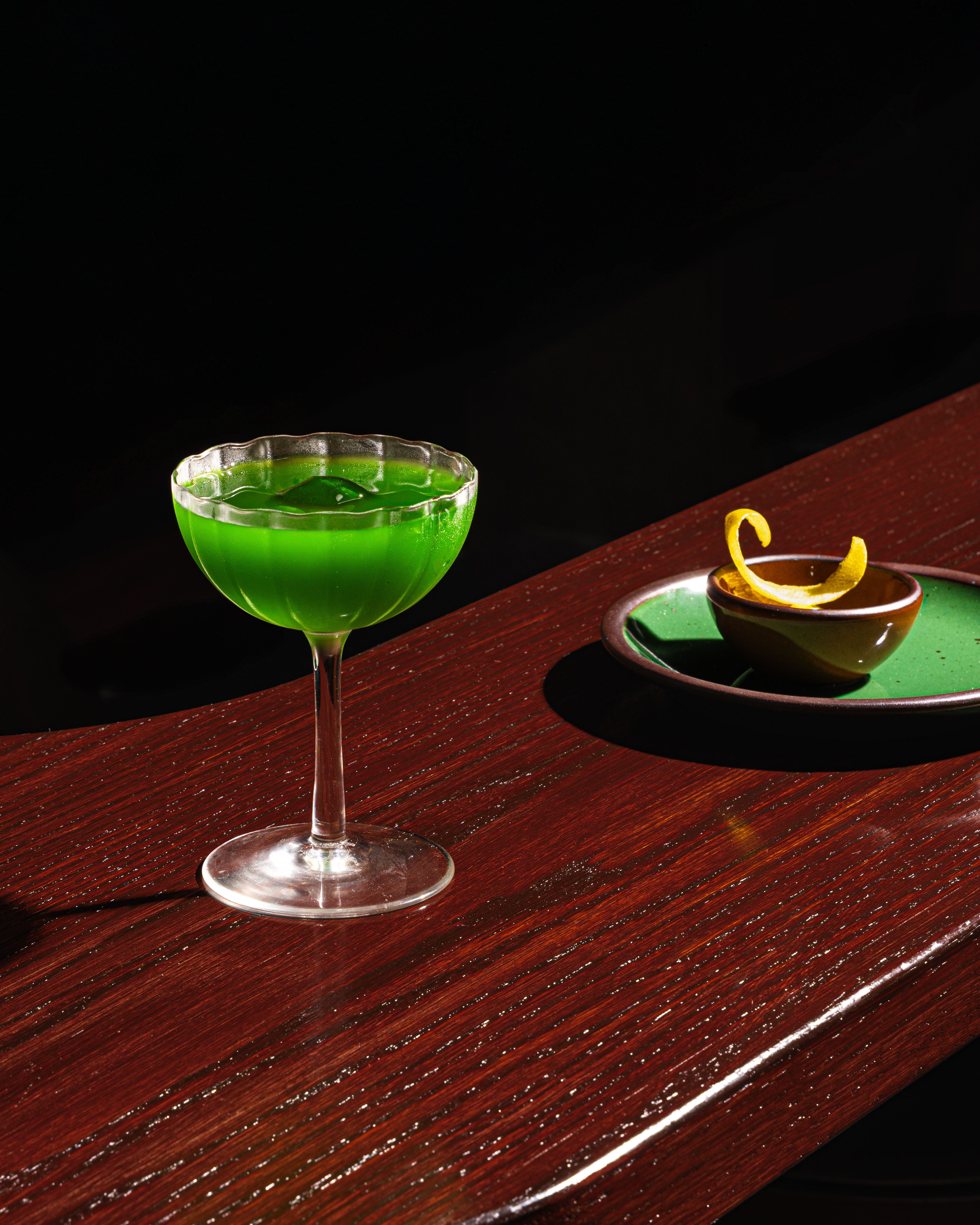 A green cocktail in a stemmed glass sits on a dark wooden counter next to a matching green plate and brown bowl holding a lemon garnish.