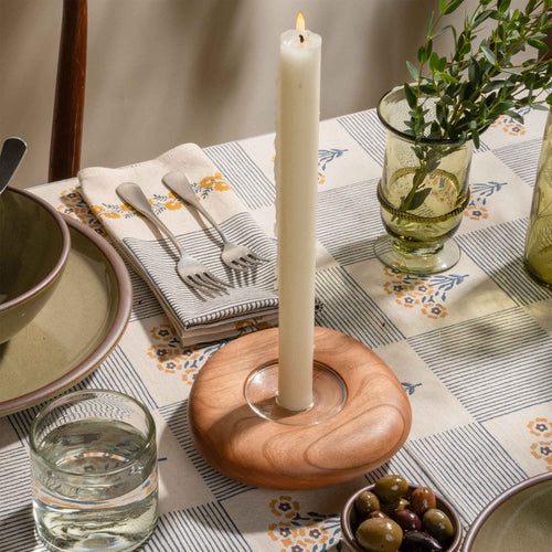 Lit white taper candle in a wooden holder on a patterned table setting with glassware and olives.