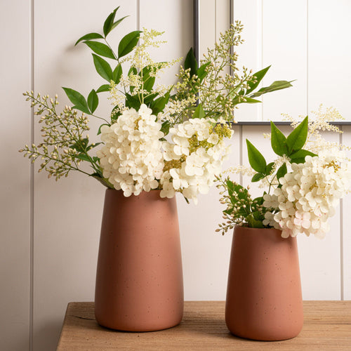Two terracotta vases with white hydrangeas and green foliage on a wooden table in front of a mirror.