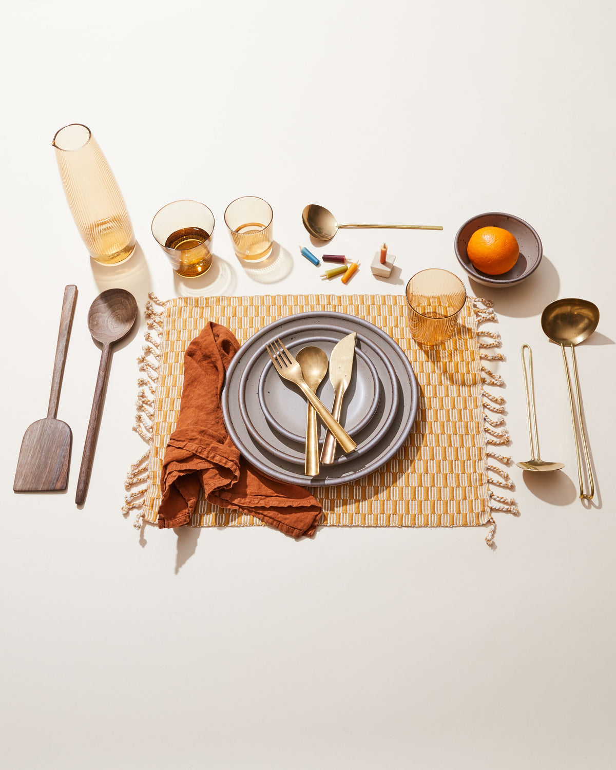 A table setting with a warm placemat, brass flatware, a terracotta napkin, and plates in a warm pale brown, and more.