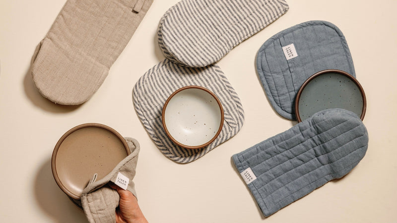 Ceramic plates in neutral tones are paired with striped and solid-colored oven mitts and pot holders on a light surface. A hand holds one plate using a beige oven mitt.