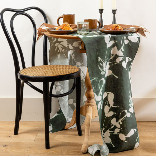 Small round table with dark green floral runner, two mugs, plates with pastries, and a black chair on wooden floor.